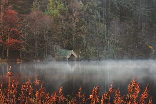A misty autumn morning at Loch Ard with a serene boathouse and reflections, surrounded by fall foliage in the Scottish Highlands.