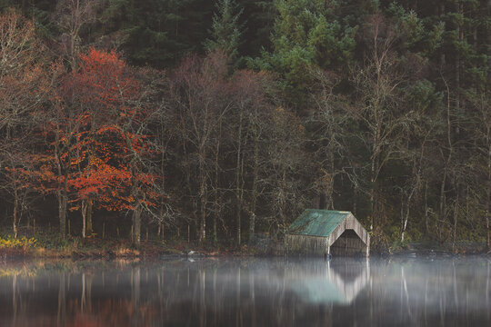 A misty autumn morning at Loch Ard with a serene boathouse and reflections, surrounded by fall foliage in the Scottish Highlands. - Powered by Adobe