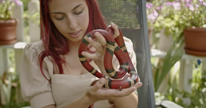A young woman with red hair sits on a garden chair and plays with her pet snake.