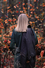 A blonde girl stands with her back in the autumn forest. Leather jacket on the shoulder.