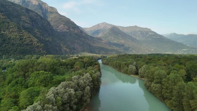 A drone pans down over the Toce River, nestled between trees, with majestic mountains and a clear sky in the background, creating a breathtaking view of nature in Ansola, Italy