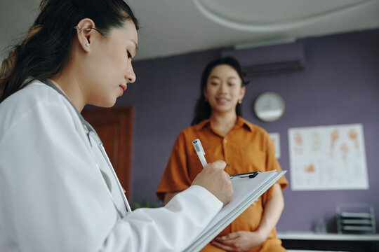 Pregnant woman with Asian descent consults with doctor in clinic. Medical professional wearing white coat holds clipboard while patient listens attentively in chair