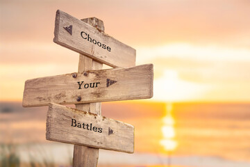 choose your battles text quote written on wooden signpost by the sea during sunset and golden hour.