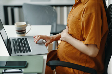 Expectant mother sitting at desk, engaged with laptop, holding belly gently, coffee mug nearby, embodying modern working lifestyle and balance