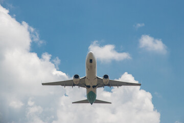 A commercial passenger plane soars through a blue sky, flanked by soft white clouds floating in the background.