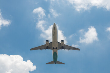 A commercial passenger plane soars through a blue sky, flanked by soft white clouds floating in the background.