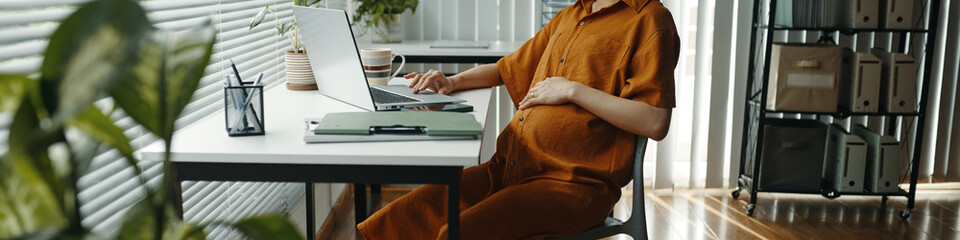 Pregnant woman sitting at desk in contemporary office, focused on writing notes beside laptop....