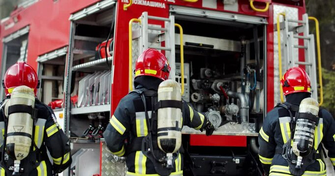 Rear of firefighters in full equipped costumes with water balloons and helmets running to water truck and working fast in black smoke. Back view on watermen coming to emergency fire place.