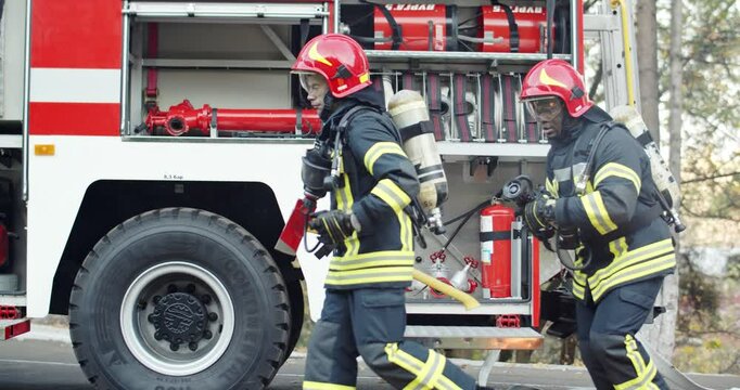 Brave multiethnic firefighters in full equipment and helmets running to fight with fire outdoor. Open fire and back smoke. Red water truck. Mixed-races courageous firemen working.