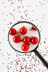 Red Christmas ornament balls under a magnifying glass over glittering sequins on a white background. Minimal Christmas holiday or Happy New Year concept. Close up.