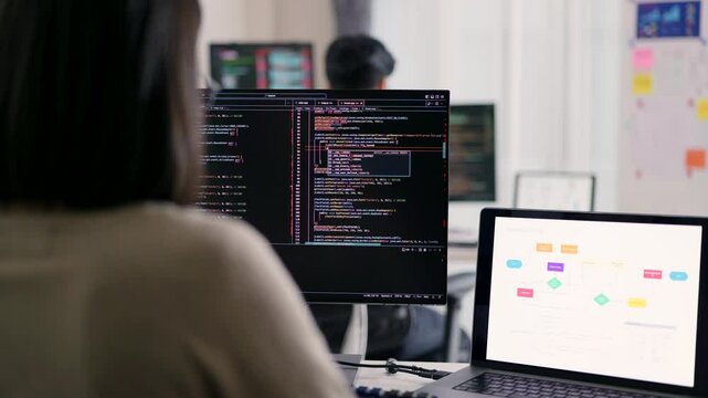 A female developer is observed from behind, scrutinizing a complex codebase on her monitor within a modern technological workspace, indicative of her problem-solving capabilities.