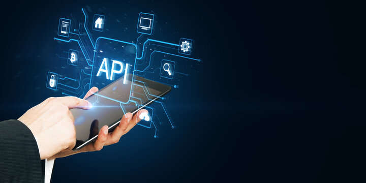 Close up of businessman hand pointing at tablet with glowing api data hologram on blue background with mock up place. Big data, Application programming interface concept.