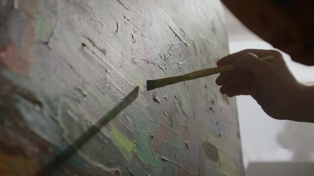 Low-angle closeup of hand of anonymous artist adding fine details and colour accents to abstract oil painting, while drawing on easel in workshop, against window, in hazy day light