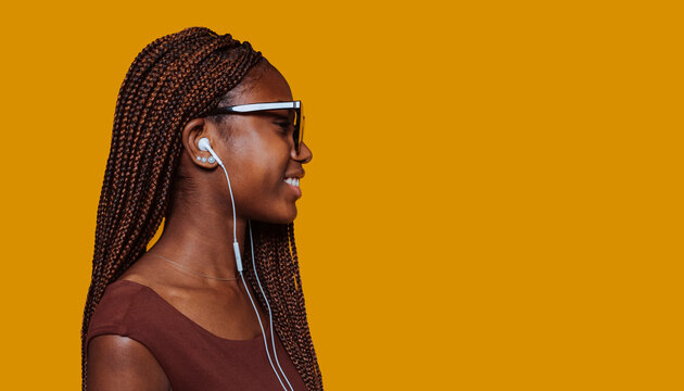 Side view of young african girl in casual brown top and sunglasses, looking away, listening music with earphones, broadly smiles isolated over yellow background at studio.