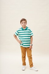 Vertical shot of little boy wearing smartwatch posing with hand on waist while showcasing casual outfit of striped green polo shirt and yellow trousers on neutral background in studio, copy space