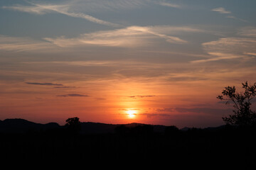sole che tramonta dietro boschi e colline in un ambiente rurale e naturale di campagna e colora di arancione e rosso il cielo coperto da nuvole e da una foschia leggera