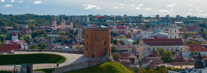 Obraz premium Gediminas' Tower stands on a hill in Vilnius, Lithuania, surrounded by greenery. The cityscape features modern and historical architecture under a cloudy sky.