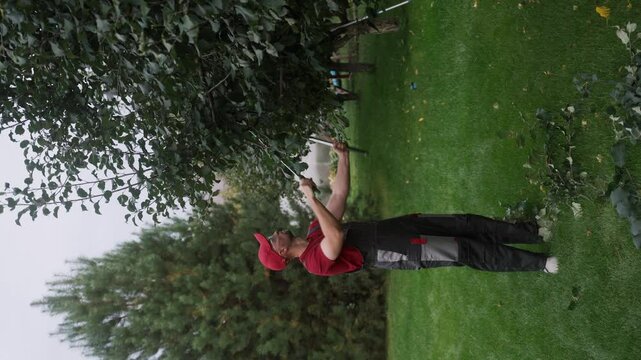 Male gardener cuts a tree branch using long loppers while wearing work clothes and a cap. Focused on the task, he demonstrates skill and precision in maintaining the garden