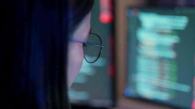 An attentive female developer with glasses analyzing source code on a computer monitor in a modern office at night.
