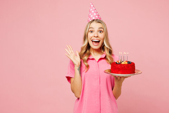 Young shocked surprised happy woman she wear shirt cone hat celebrating birthday holiday and hold cake with candles spread hand look camera isolated on plain pastel pink background. Lifestyle concept.