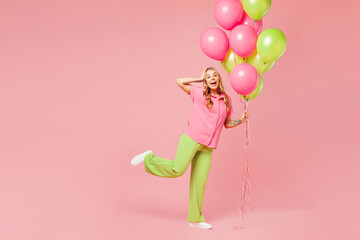 Full body side view young surprised woman wear shirt celebrating birthday holiday stand hold bunch of colorful air balloons raise up leg pov fly isolated on plain pink background. Lifestyle concept.