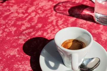 dettaglio di una tazzina bianca riempita con del caffè espresso caldo su un tavolo coperto da una tovaglia rossa, all'aperto, illuminato dal sole, di mattina