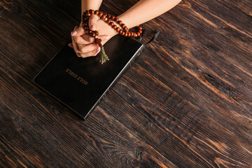 Hands of praying woman with rosary beads and Holy Bible on wooden background