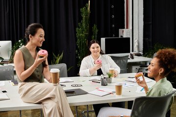 Three professional women share a lighthearted moment over coffee and donuts in an office setting.