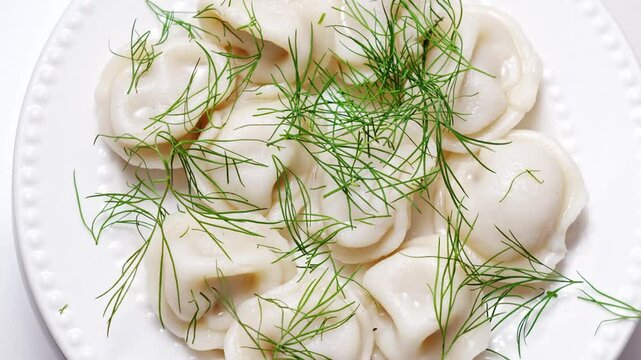 revolves. Boiled dumplings with dill are arranged in a circle on a white plate. The plate with dumplings is set against a light background, creating a simple and appetizing composition.