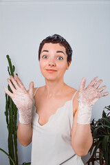 A short-haired woman in a light-colored top wears latex gloves and holds up her hands with a humorous expression, as if caught in a messy situation. Cacti and plants are visible in the background.