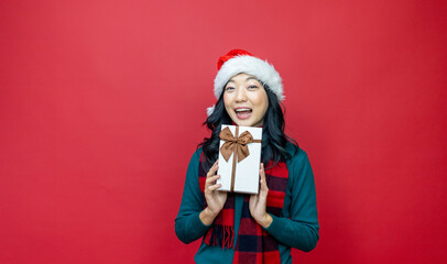 Pretty smiling asian woman in warm christmas sweater and santa hat holding gift box as present with red background for season celebration