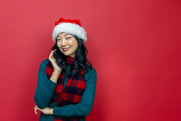 Pretty smiling asian woman in warm christmas sweater and santa hat on red background for season celebration