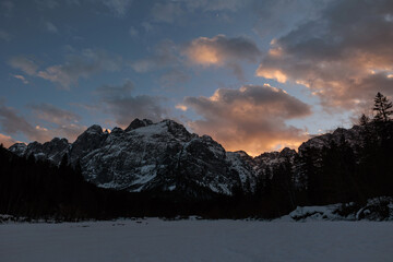 sole che tramonta dietro a delle alte montagne innevate, in un cielo azzurro e parzialmente...