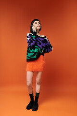 A young woman poses confidently in a colorful sweater, showcasing her tattoos against a vivid orange background.