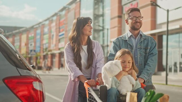 Young family went shopping. Smiling husband and wife push cart with groceries and their daughter. Happy little girl ride in cart and holding soft toy. Couple in love are talking and walking to car.