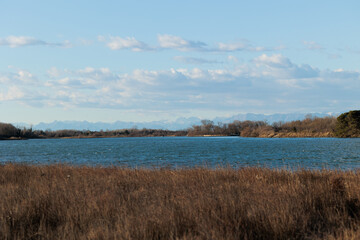 guardando da una delle sponde di un ramo del fiume Isonzo verso la pianura e le montagne distanti della regione Friuli Venezia Giulia, di giorno, con cielo parzialmente sereno, in inverno