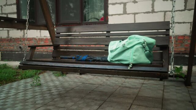 Mint-colored bag and blue umbrella resting on wooden swing bench outside, with background showing partially constructed building, greenery, and interlocked pavement