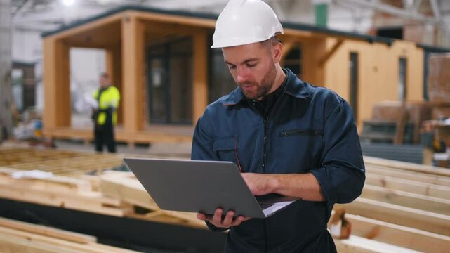 Laptop in hands. Male builder is at construction of wooden house.