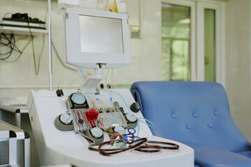 Medical tool with tubes and monitor placed next to a blue chair in hospital room with white walls and shelves holding devices in the background