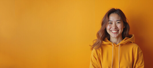 Cheerful Asian woman in yellow hoodie smiling against orange background with copy space