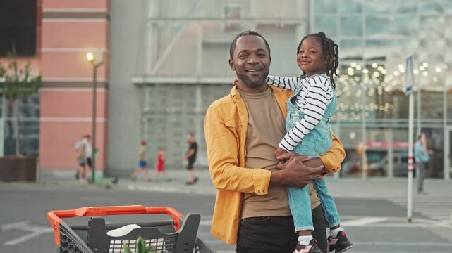 Smiling African American dad and daughter pose for camera. Dad holds daughter in arms and looks at camera. Girl is satisfied and huggs dad by neck. Dad and daughter are standing near grocery cart.