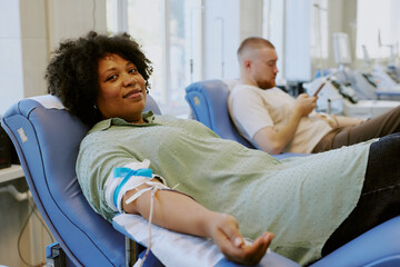 Smiling individual donating blood while sitting on comfortable chair in medical facility, another...