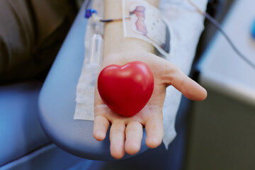 Close-up image showing person holding heart-shaped stress ball while donating blood. IV line visible on arm with background slightly blurred, emphasizing action