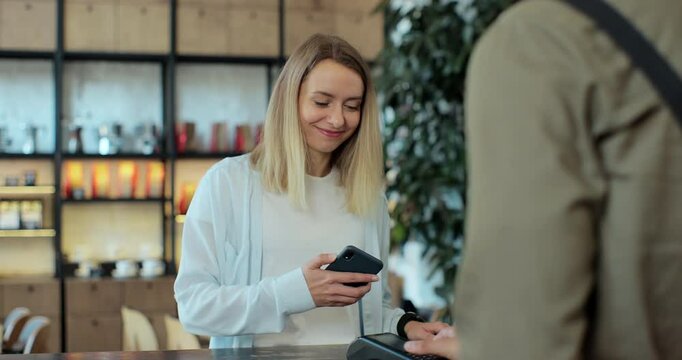 In the cafe barista man makes takeaway coffee for a woman customer which pays by contactless mobile phone to credit card system.