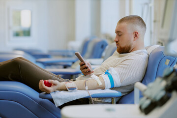 Caucasian man sitting in a comfortable chair with arm extended, donating blood while using...