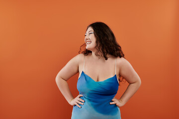 Obraz premium A young plus size woman showcases her style in a blue dress, smiling joyfully with an orange backdrop.