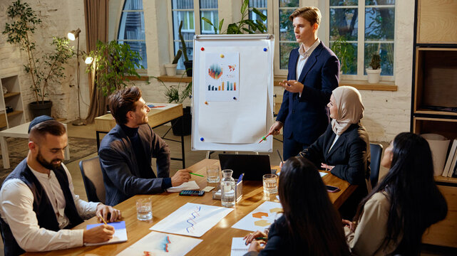 Young man explaining global market data to diverse team, using flipchart to present detailed charts and trends in corporate meeting setting. Concept of dei in business, teamwork