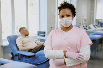 Healthcare worker in pink uniform standing in front of patient seated for blood donation in a medical facility equipped with necessary equipment and beds