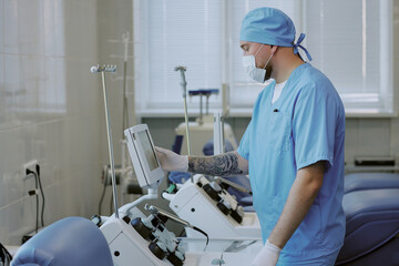Fototapeta premium Medical professional in blue scrubs and surgical cap adjusting equipment in hospital room, with intravenous drip stands and medical devices around, focusing on patient care
