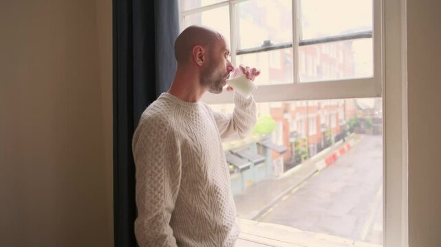 slender young man drinks fermented milk product kefir from glass by window, looking out at street, bustling cityscape, healthy lifestyle choice, mindful moment self-care, moment of calm